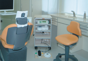 Behandlungszimmer mit orange-grauem Behandlungsstuhl, Hocker und Rollwagen mit Geräten, davor ein Fußschalter