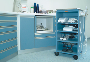 Praxisraum mit blauen Schränken und passendem blauem Rollwagen mit offenen Fächern, Geräten und Schläuchen